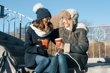 Two young teenage girls having fun outdoors, girlfriends in winter clothes, positive people and friendship concept. Girls drinking hot drinks and talking on a sunny winter day