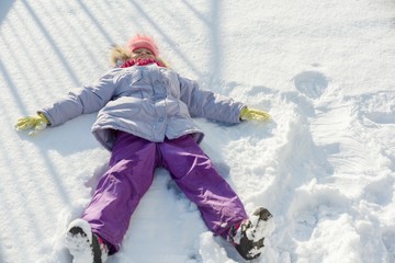 Fototapeta na wymiar Winter portrait of cheerful child girl having fun in the snow, lying on the snow in the form of angel, top view