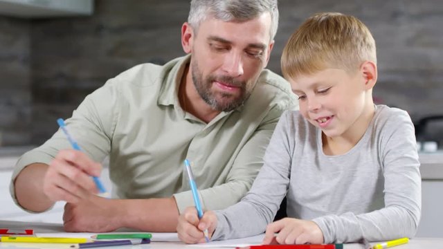 Mid-aged bearded father smiling and talking with little son while drawing together with colored pencils at kitchen table