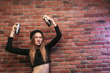 Image of aggressive hip hop girl 20s, standing against brick wall with spray cans