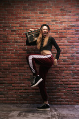 Full length image of teenage woman 20s, standing against brick wall and holding boombox