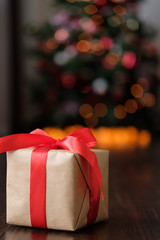 A gift in craft paper with a red ribbon on the background of a New Year's decorated Christmas tree with lights