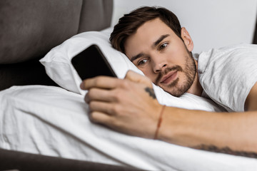 handsome bearded young man lying in bed and using smartphone