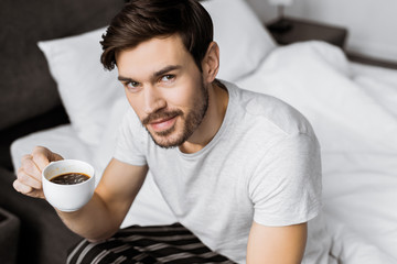 handsome young man holding cup of coffee and smiling at camera while sitting on bed in the morning