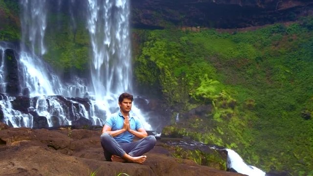 strong guy sits in pranayama yoga pose on rock at waterfall