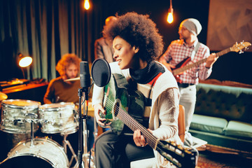 Mixed race woman singing and playing guitar while sitting on chair with legs crossed. In background drummer and bass guitarist. - obrazy, fototapety, plakaty