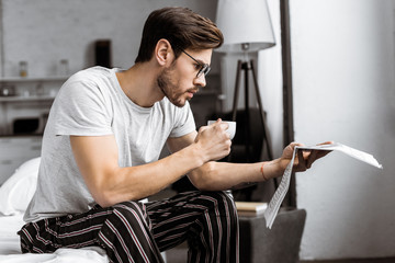 handsome young man in pajamas and eyeglasses holding cup of coffee and reading newspaper in the...