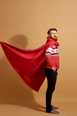 Red-haired man with beard dressed in a red and white sweater with deer and red cape  fluttering in the wind and sleepers stands on a beige background
