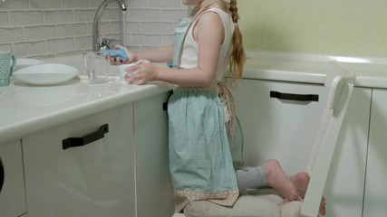 beautiful little girl in an apron is standing on a chair, in the bright kitchen, doing the dishes with a sponge, helping parents