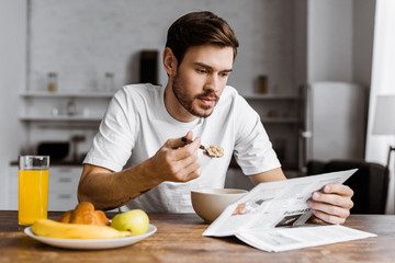 handsome young man having cereal for breakfast and reading newspaper at home - obrazy, fototapety, plakaty