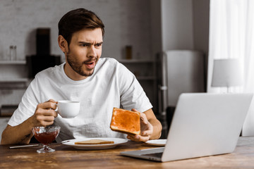confused young freelancer eating toast with jam and looking at laptop screen at home
