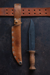 Combat soldier Stainless steel fighting knife with leather handle, scabbard and leather belt in  wooden vintage frame. Close-up, top view on dark brown concrete background.