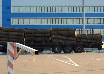 Naklejka na ściany i meble Lumber trailer loaded wooden logs on the asphalt road in front of cargo building