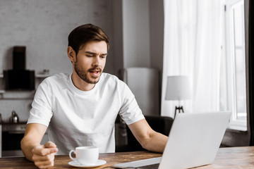confused young freelancer with coffee using laptop at home