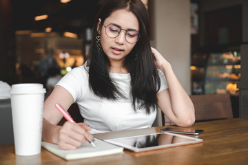 Beautiful asian business woman wearing glasses is working and writing on notebook in cafe.