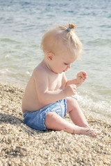 Candid photo of adorable blonde baby on the pebble beach near the sea, summer vacation on the seaside