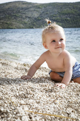 Fototapeta na wymiar Candid photo of adorable blonde baby on the pebble beach near the sea, summer vacation on the seaside