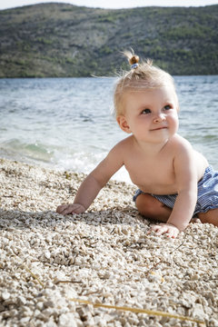 Candid photo of adorable blonde baby on the pebble beach near the sea, summer vacation on the seaside