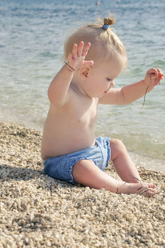 Candid photo of adorable blonde baby on the pebble beach near the sea, summer vacation on the seaside