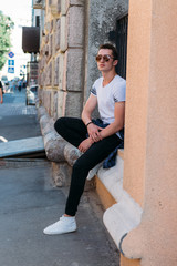Attractive, sexy guy sits on the street in sunglasses. self-confidence, flatulence and arrogance. model, posing, portrait of a young man in white sneakers