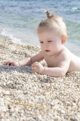 Fototapeta na wymiar Candid photo of adorable blonde baby on the pebble beach near the sea, summer vacation on the seaside