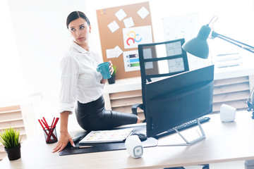 Obraz premium A young girl sat down on a table in the office, holding a Cup and working with a computer and documents.