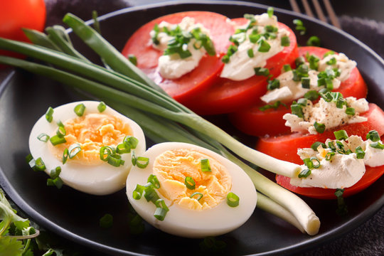 Half boiled eggs with tomatoes, green onions, sprinkled with greens in a black plate on a gray wooden table. Close-up