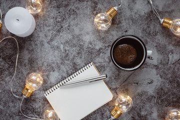 Winter cozy concept. Candle and coffee drink, notebook, pen and decorated with led lights on gray table.