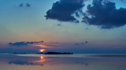 Obraz premium Wide Shot Of Red Setting Sun Through Blue Dark Clouds Over A Small Atoll Island In The Calm Sea Of The Maldives.