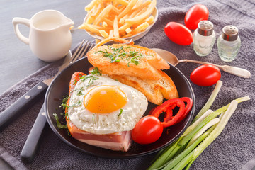 Fried egg with bacon in a black plate with fried pieces of bread, greens tomatoes, a jug of milk and French fries on a gray wooden table. Close-up