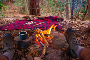 First person view of man relaxing / having tea in an enamel outdoor mug by a campfire in Blue Ridge / Appalachian Mountains trail near Asheville, North Carolina. Boots, Bushcraft, blanket, Axe.