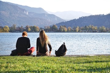 Two best friends sitting and relaxing on the grass field front of the beautiful lake and mountain background.Chilling out on a good time.Summer vacation, happy holidays, travel and people concept.