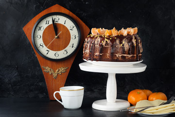 Dessert cake with chocolate icing decorated with tangerines on a black background. Clock wooden