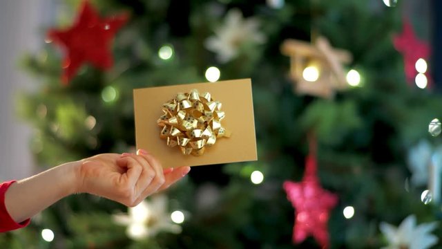 Closeup on housewife near Christmas tree showing Christmas gift card. Xmas it's the most wonderful time of year! What secrets does a golden gift card hold?. This video was shot in PRORes 422 codec.