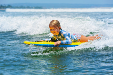 Happy baby girl - young surfer ride on surfboard with fun on sea waves. Active family lifestyle, kids outdoor water sport lessons and swimming activity in surf camp. Beach summer vacation with child. - obrazy, fototapety, plakaty