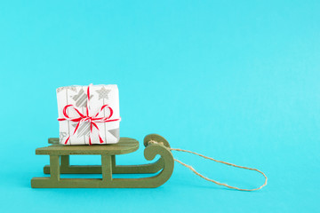 Small gift box wrapped of white and gray paper with red-white ribbon on a green wooden sleigh on a bright blue background. Christmas and New Year concept with copy space.