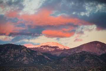 Fototapeta premium Dramatyczny, piękny zachód słońca rzuca fioletowe i pomarańczowe kolory i odcienie na chmury i ośnieżony szczyt Santa Fe Baldy w górach Sangre de Cristo w pobliżu Santa Fe w Nowym Meksyku