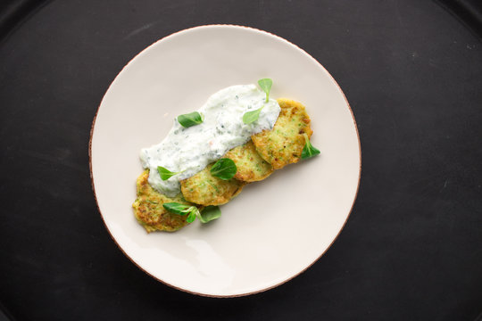 Zucchini pancakes with greens and sour cream on black background