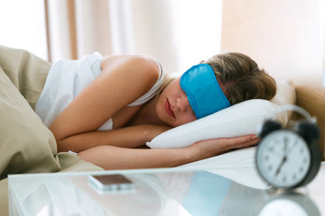 Beautiful young sleepy woman with sleep mask resting in bed with alarm clock in bedroom at home.
