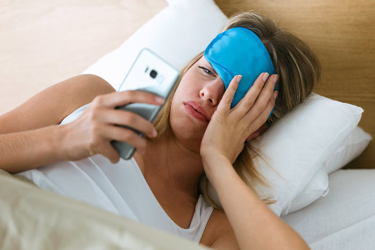 Tired young woman lying in bed pulling up sleeping mask to see what time is it at smartphone in bedroom at home.