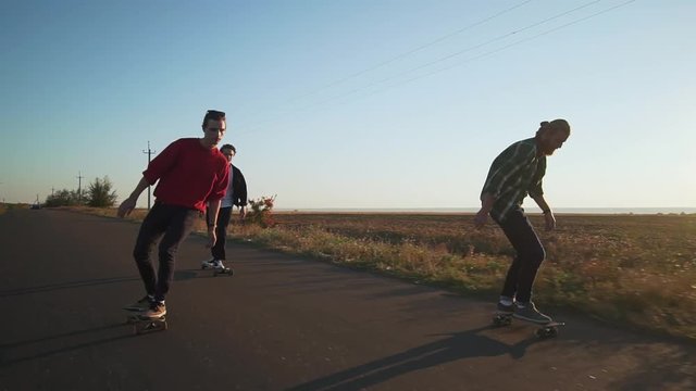 Three hipster men skateboarding at empty road at sunset rapid slow motion