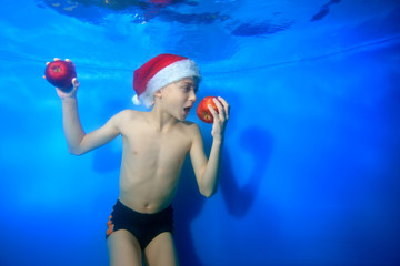 The child prepared to bite off the red Apple, which he holds in his hand. He poses underwater on a blue background in Santa's hat. Concept. Close up. Horizontal orientation