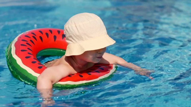 Full shot smiling little baby wearing hat floating in outdoor swimming pool enjoying vacation at summer sunny day using lifebuoy high angle European kid playing and having fun in pure clean water