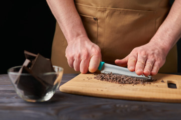 A man in a brown apron with a knife chopping chocolate