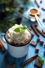 Mug with hot chocolate and whipped cream, decorated with ground cinnamon and mint leaves on a blue wooden table