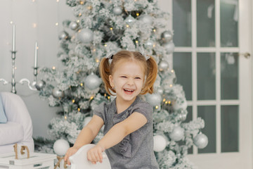 beautiful little girl in a white dress like a princess is sitting on a toy wooden horse in a vintage studio, New Year and Christmas photography.