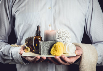 Man holding white ceramic tray with group of different spa essentials, beard oil, gray soap,...