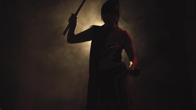 interesting video, a samurai brunette girl with a long pony tail and with two katanas in her hands is standing in a dark room with a dim light, posing and slowly turning to the camera