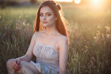 Beautiful young woman with perfect make up and long plaited hair sitting in the field at sunset. Wearing cotton ethnic summer dress. Backlight. Text space. Outdoor shot
