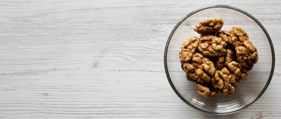 Walnuts in glass bowl over white wooden surface, overhead view. Flat lay, top view. Copy space.